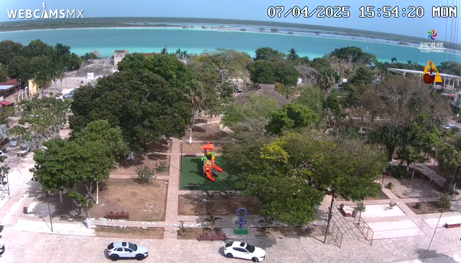 An aerial view shows a park with a playground, surrounded by lush green trees and overlooking a calm, turquoise body of water under a clear blue sky.