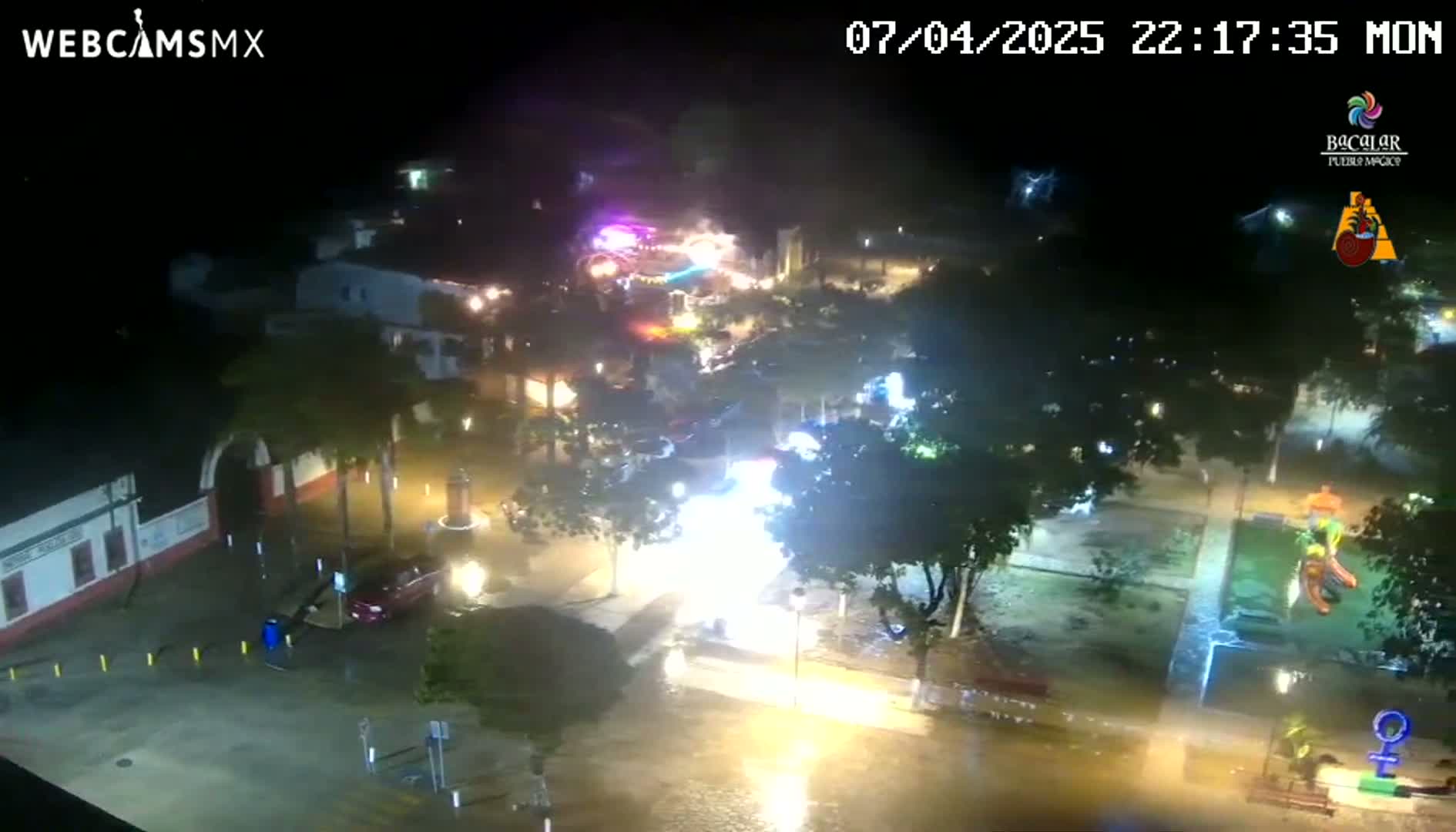 A nighttime aerial view of a flooded town square with illuminated buildings and trees, under heavy rain.