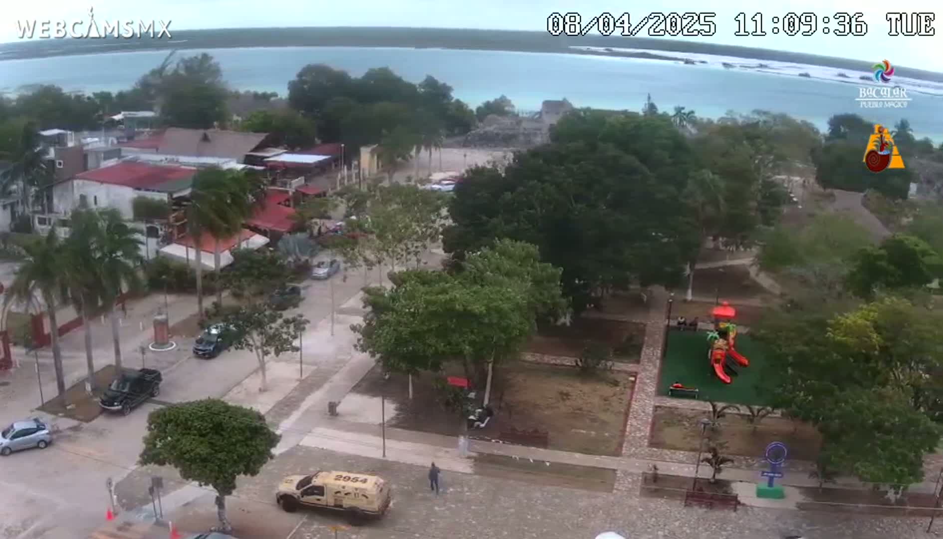 An aerial view of a town square with a playground,  vehicles parked on the streets, and a body of water visible in the background under an overcast sky.