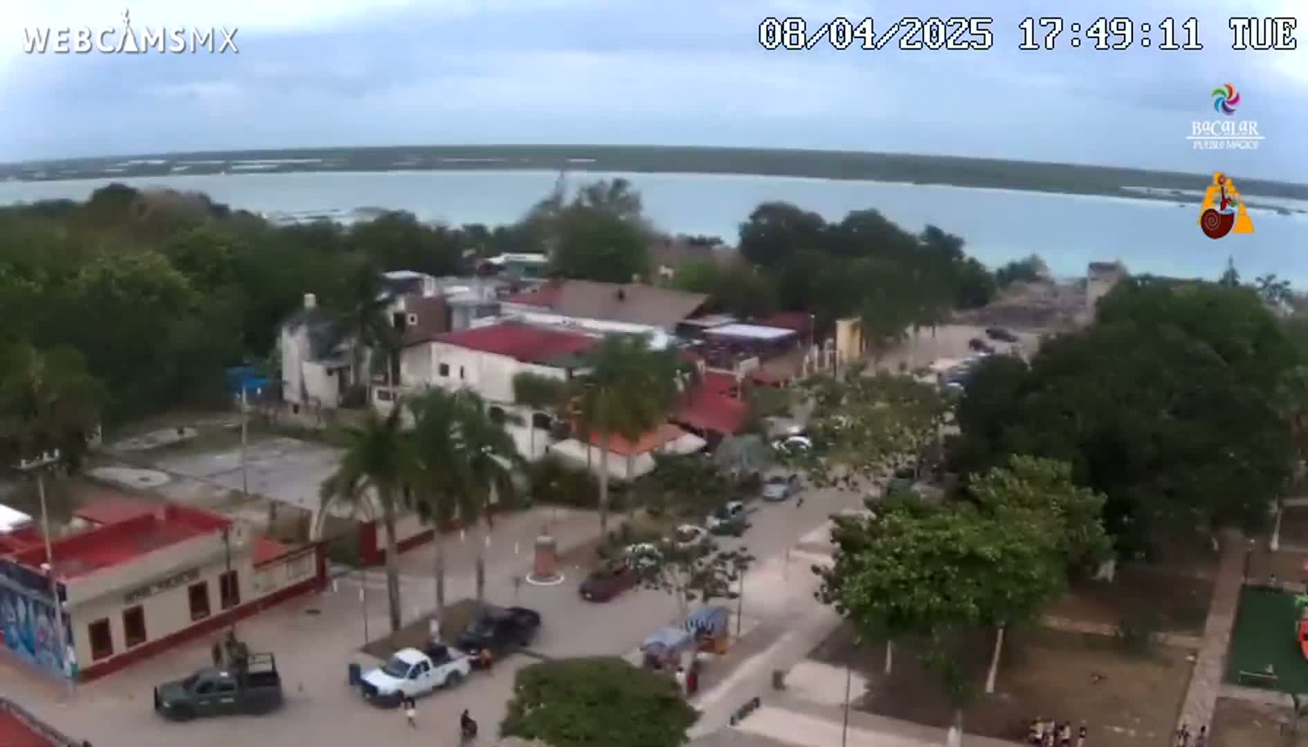 An aerial view shows a town with palm trees and buildings near a calm body of water under a partly cloudy sky.
