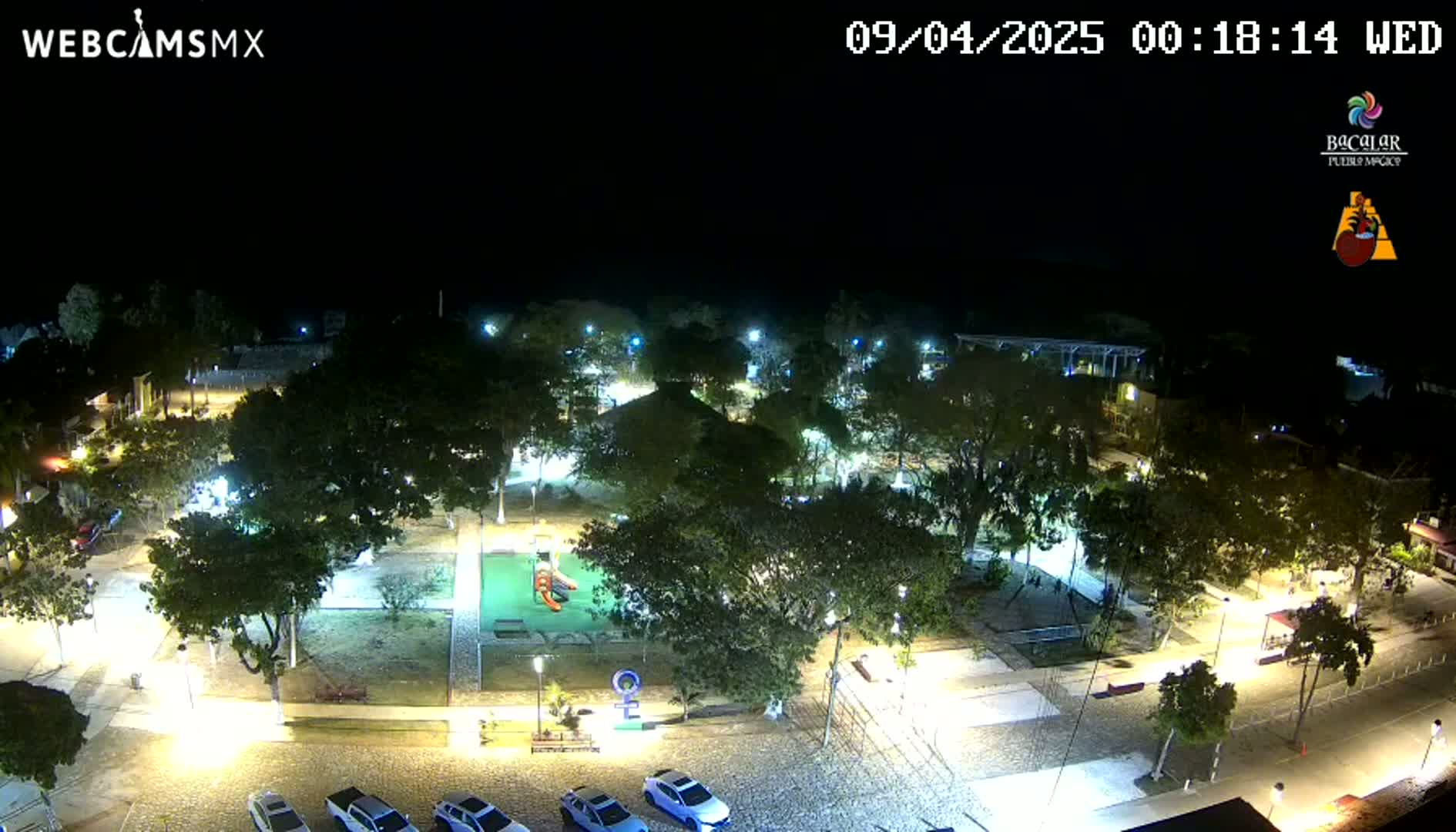 A nighttime aerial view of a plaza with trees, a playground, and parked cars under clear skies.