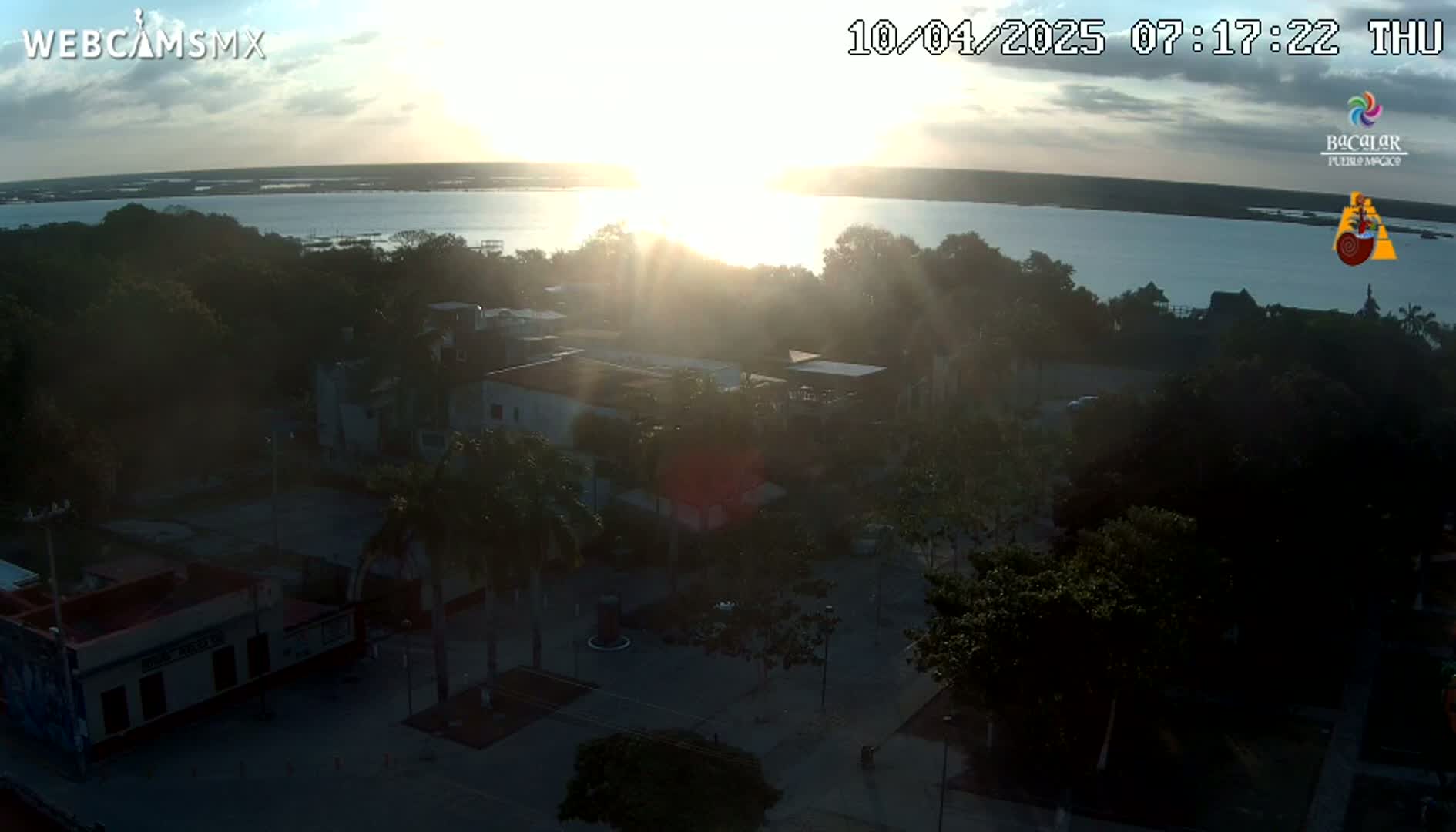 A sunrise over a calm body of water is visible from an elevated vantage point overlooking a town square with buildings and lush green trees, under a partly cloudy sky.