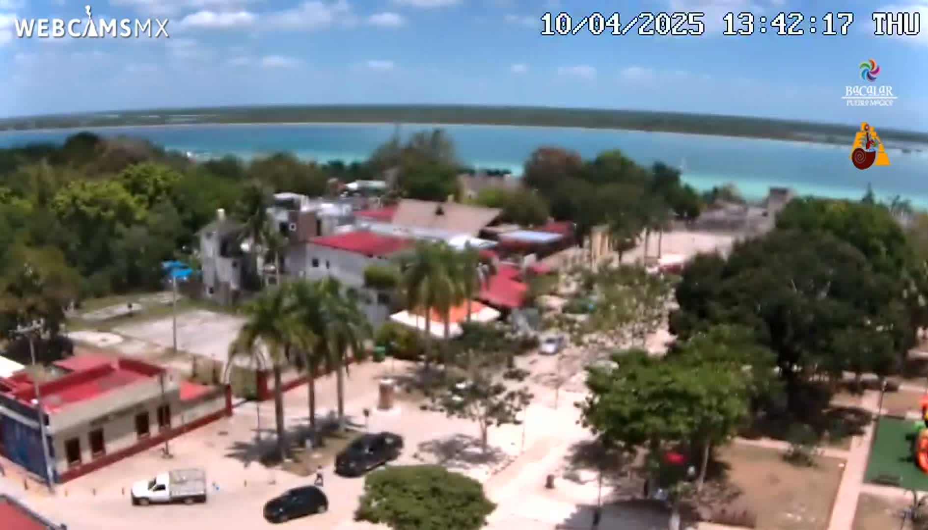 An aerial view shows a town square with buildings, palm trees, and parked cars, next to a calm, turquoise body of water under a partly cloudy sky.
