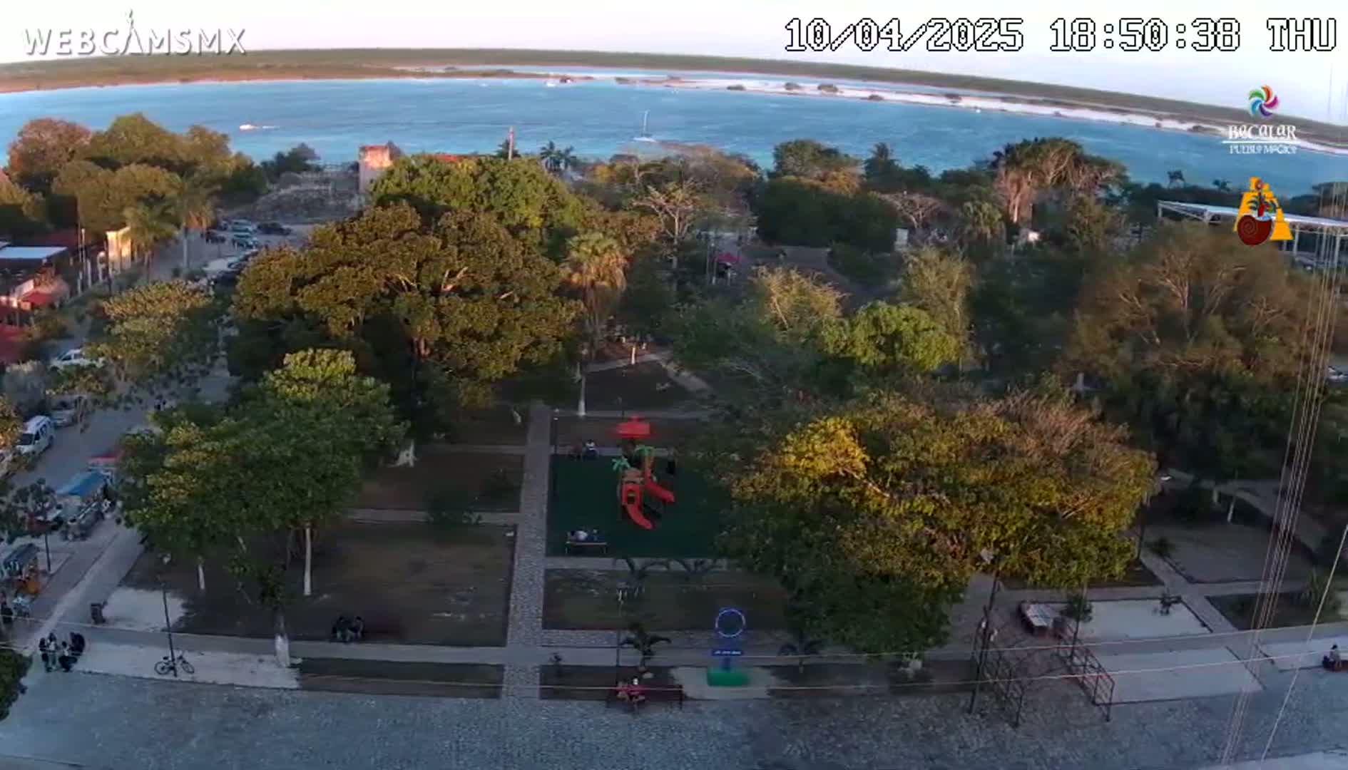 An aerial view shows a town square with a playground, surrounded by lush green trees and paved pathways, next to a calm body of water under a clear evening sky.