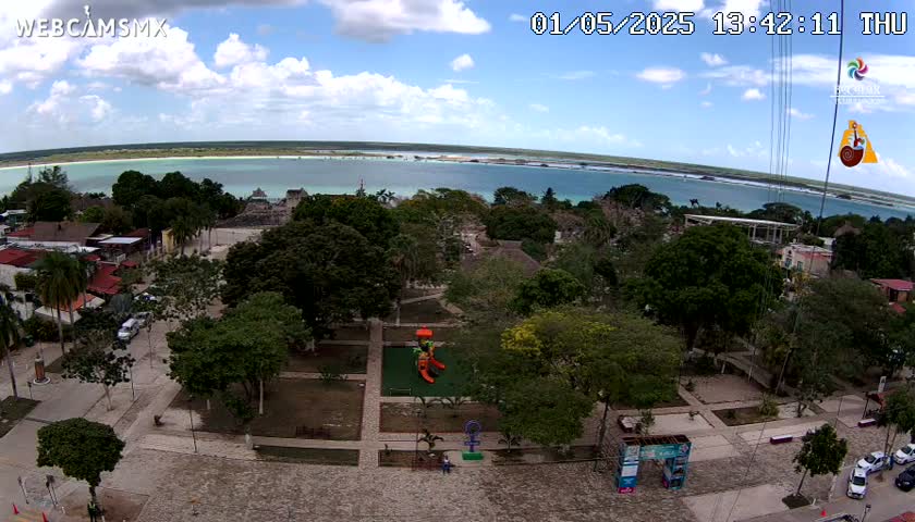 An aerial view of a town square with lush green trees and a playground, overlooking a calm, light-blue body of water under a partly cloudy sky.
