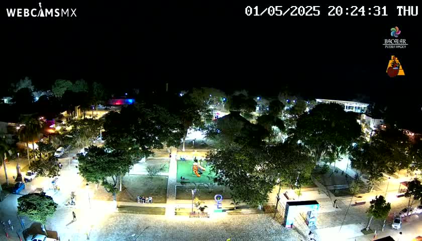 A brightly lit town square at night, filled with trees and featuring a playground, is shown under a clear, dark sky.