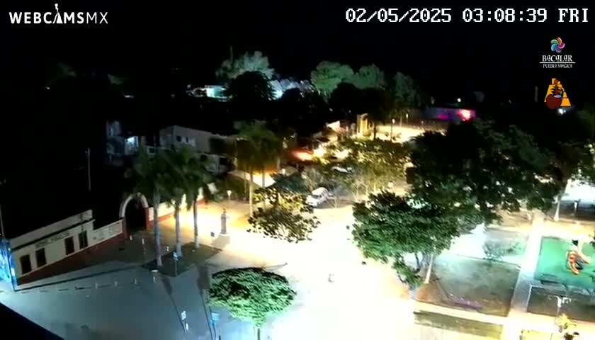An aerial nighttime view shows a town square illuminated by streetlights, surrounded by palm trees and buildings.