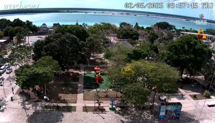 An aerial view shows a park with a playground, surrounded by trees and buildings, next to a calm, turquoise body of water under a sunny sky.