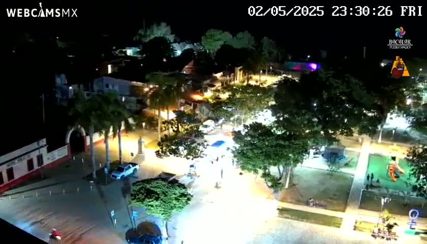 A nighttime aerial view of a town square shows buildings, trees, and a few people, with some areas appearing to be flooded under lights.