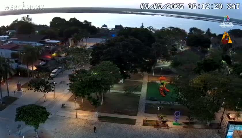 An aerial view at dusk shows a town square with a playground, trees, and buildings surrounding a paved area, next to a calm body of water.