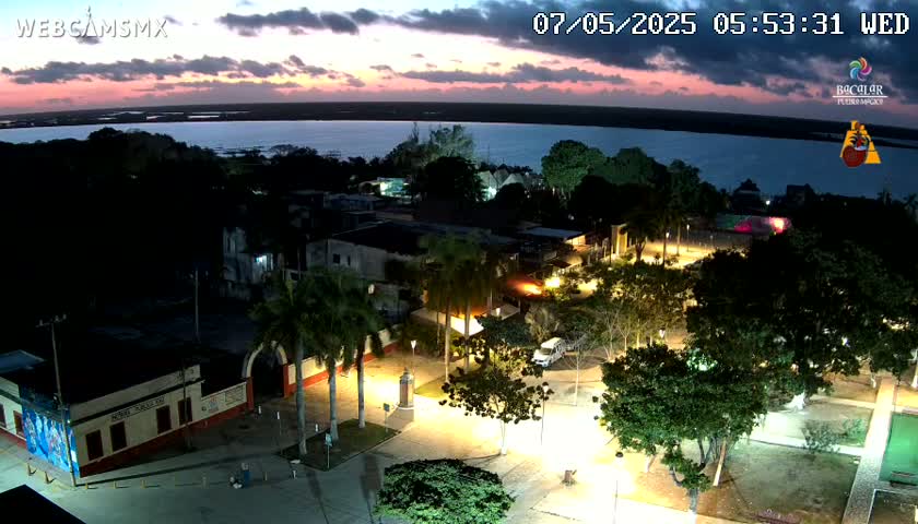 A town square at night, illuminated by streetlights, overlooks a calm body of water with a colorful sunset in the background.