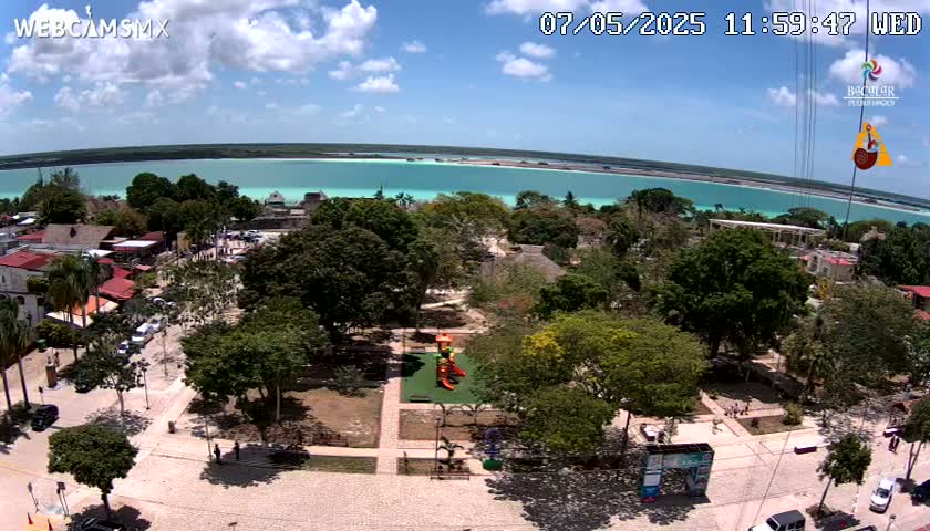 An aerial view shows a town square with lush green trees, a playground, and a turquoise body of water in the background under a partly cloudy sky.
