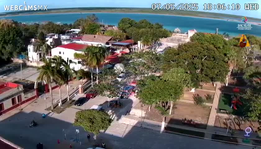 An aerial view shows a town square with buildings, palm trees, and a park, next to a body of turquoise water under a sunny sky.