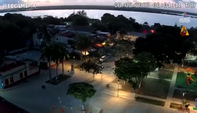 A nighttime aerial view of a town square with palm trees, buildings, and a playground, overlooking a calm body of water under a twilight sky.
