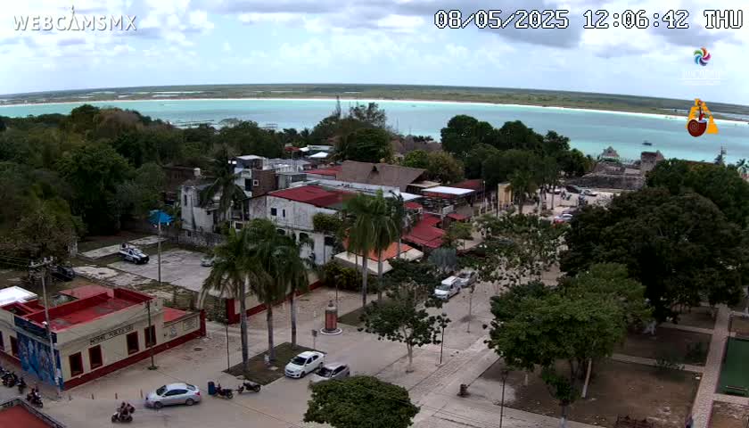 An aerial view shows a town square with palm trees, low buildings, and parked cars, overlooking a calm, light-blue body of water under a partly cloudy sky.