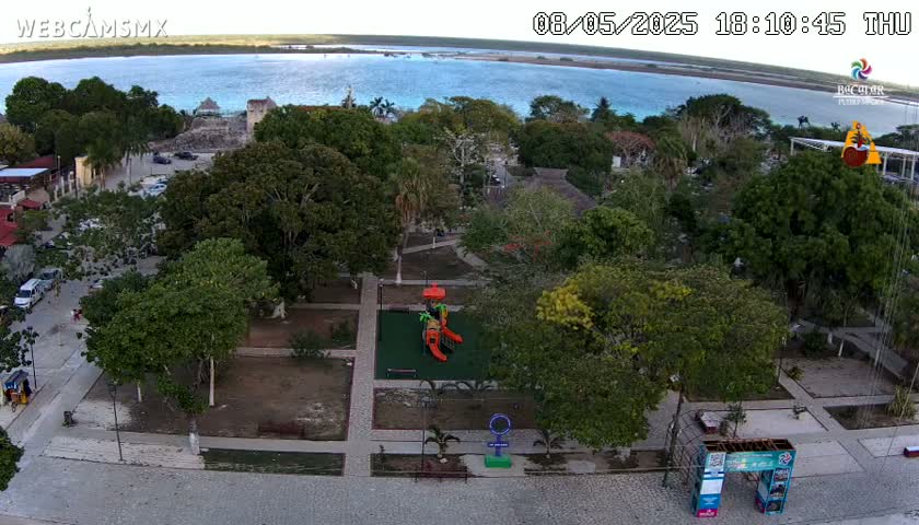 An aerial view shows a town square with a playground, numerous trees, and a calm body of water in the background under a clear sky.