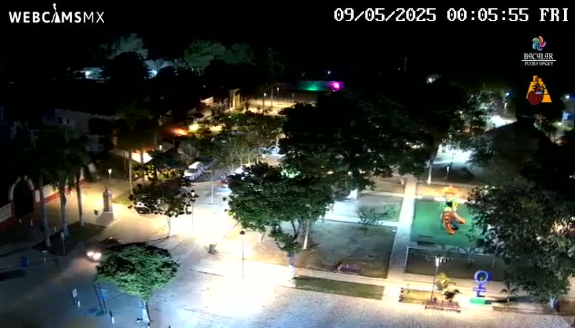 An aerial nighttime view of a town square with streetlights illuminating parked cars, trees, and playground equipment.