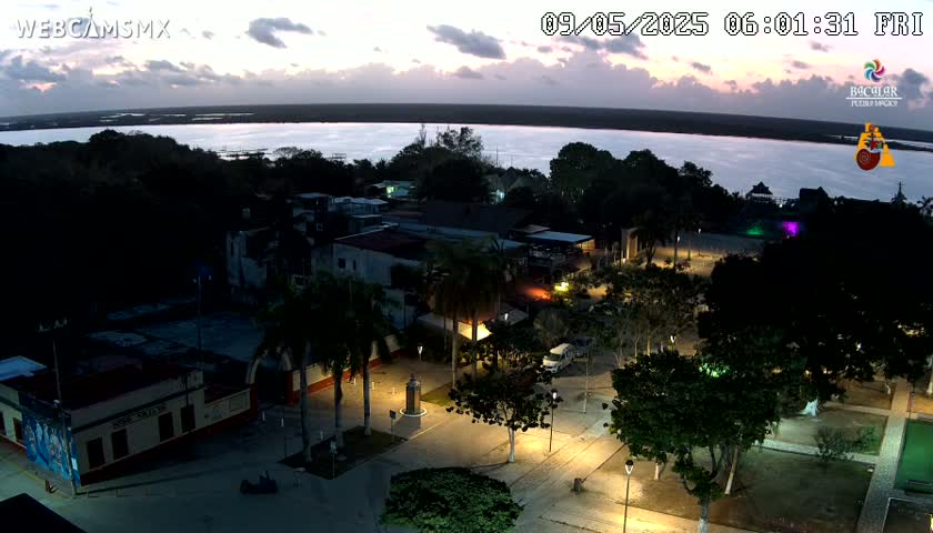 A nighttime, high-angle view of a town square illuminated by streetlights, overlooking a calm body of water under a twilight sky.