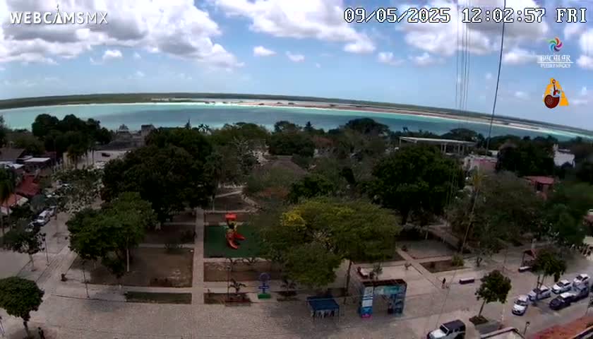 An aerial view shows a town square with a playground and surrounding buildings, overlooking a calm, light-blue body of water under a partly cloudy sky.