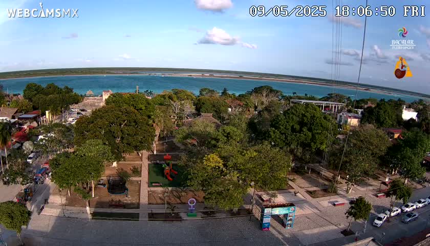An aerial view of a town square with a playground and a park, next to a body of turquoise water under a partly cloudy sky.