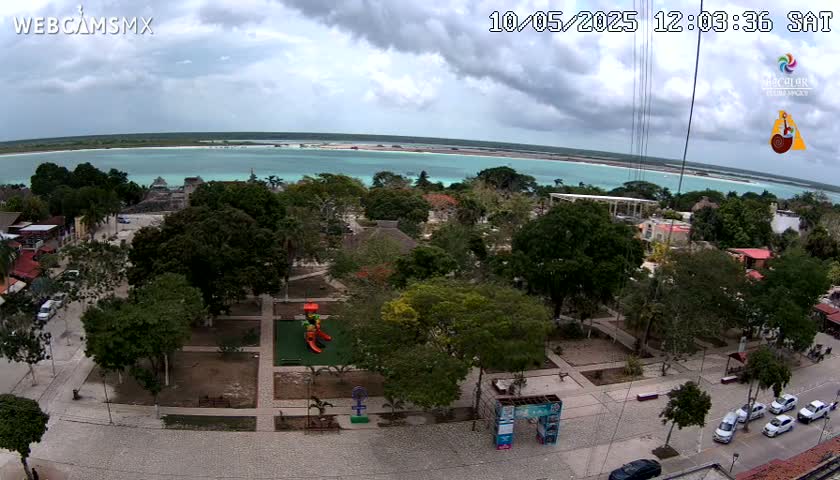 An aerial view shows a town square with lush green trees, a playground, and parked cars, overlooking a calm turquoise body of water under a partly cloudy sky.
