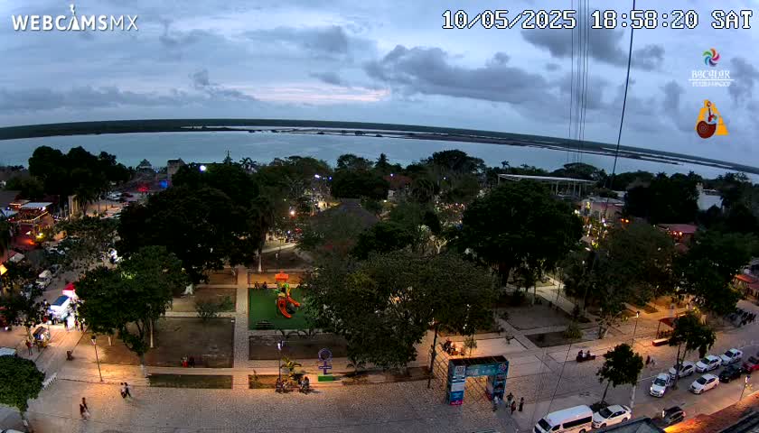 An aerial evening view of a town square with a playground, lush trees, and parked cars, overlooking a calm body of water under a partly cloudy sky.