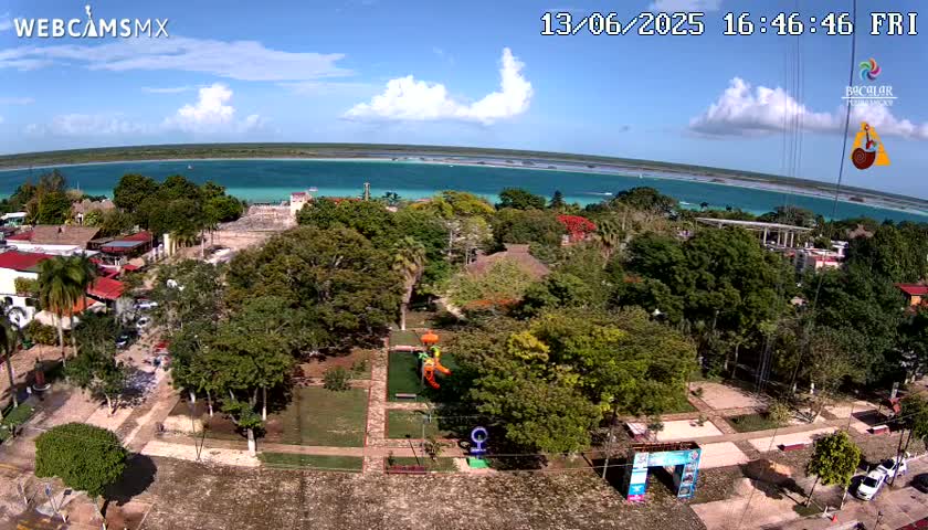 An aerial view of a town with a playground near a park overlooking a calm, turquoise body of water under a partly cloudy sky.
