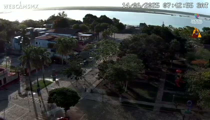 An aerial view shows a town square with buildings, palm trees, and a body of water in the background under a mostly clear sky.