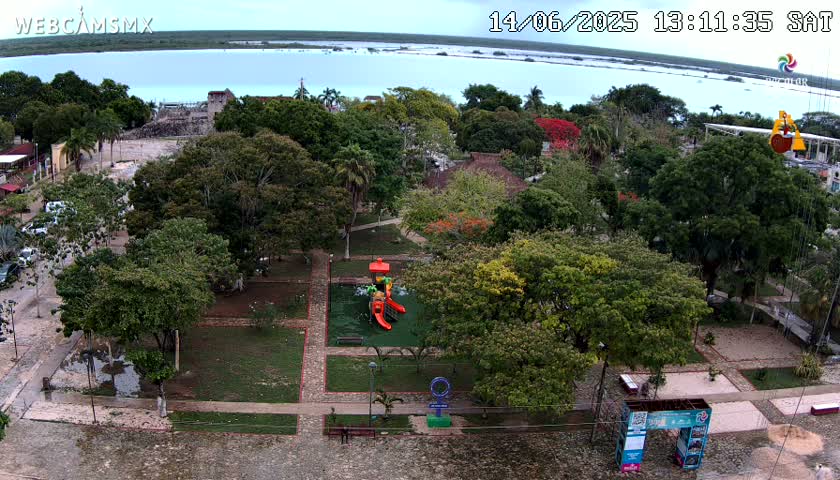 An aerial view shows a park with playground equipment, lush green trees, and a stone-paved area next to a body of calm, light blue water under an overcast sky.