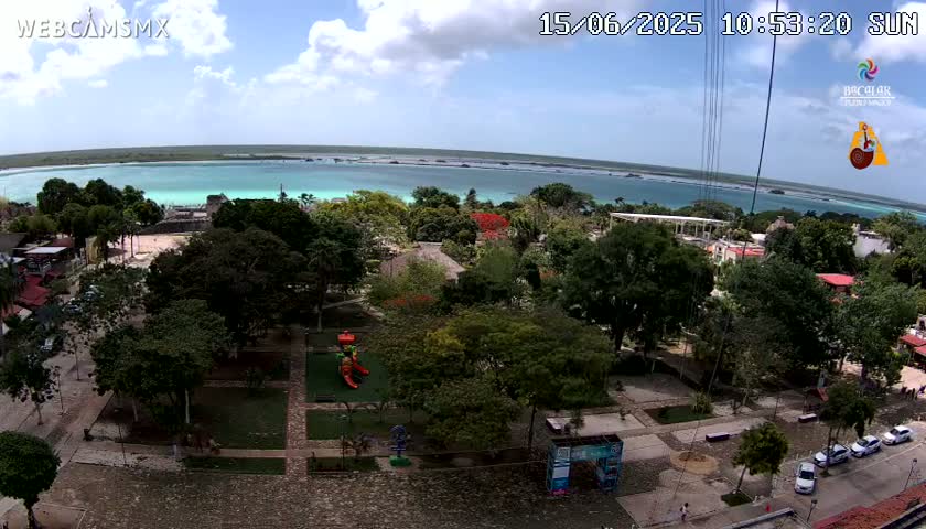 An aerial view shows a park with lush green trees and a playground, overlooking a calm, turquoise body of water under a partly cloudy sky.