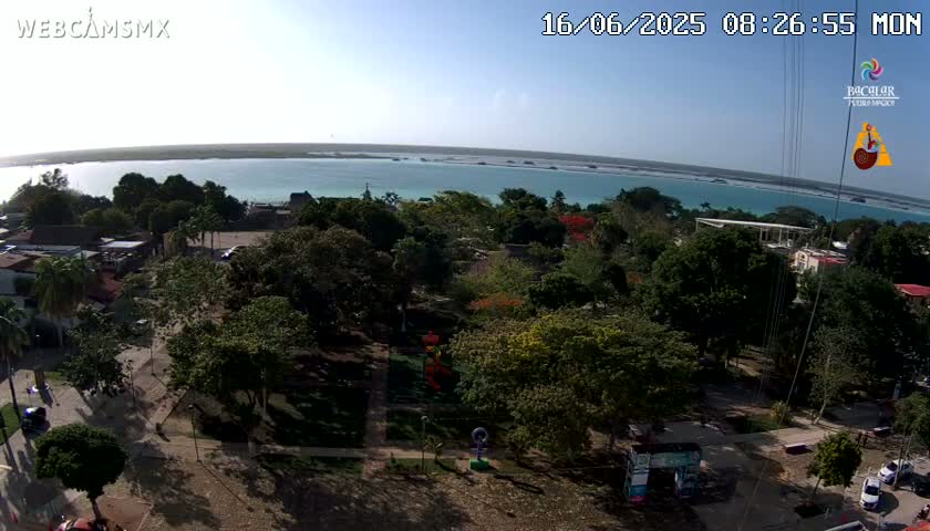 An aerial view shows a town with a park near a calm, turquoise body of water under a sunny sky.