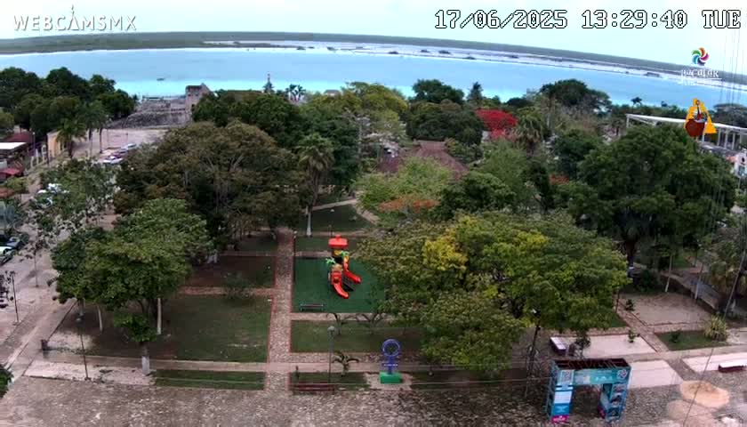 An aerial view of a park with playground equipment and a body of water visible in the background on a partly cloudy day.