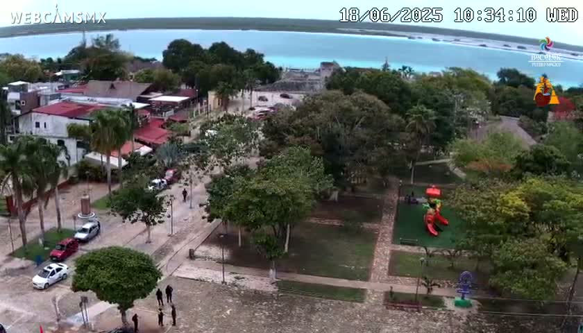 An aerial view shows a town with buildings, streets, cars, and a park with playground equipment next to a calm body of water under a partly cloudy sky.