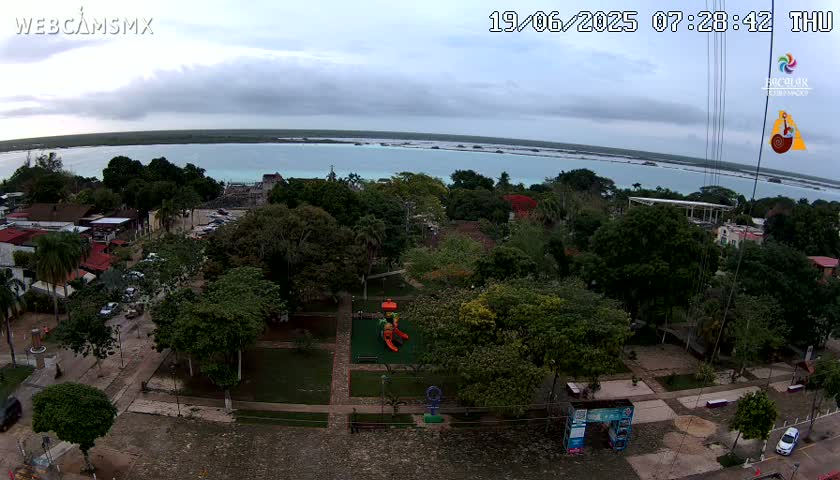 An elevated view of a town square with lush green trees and a playground, overlooking a calm turquoise body of water under a cloudy sky.
