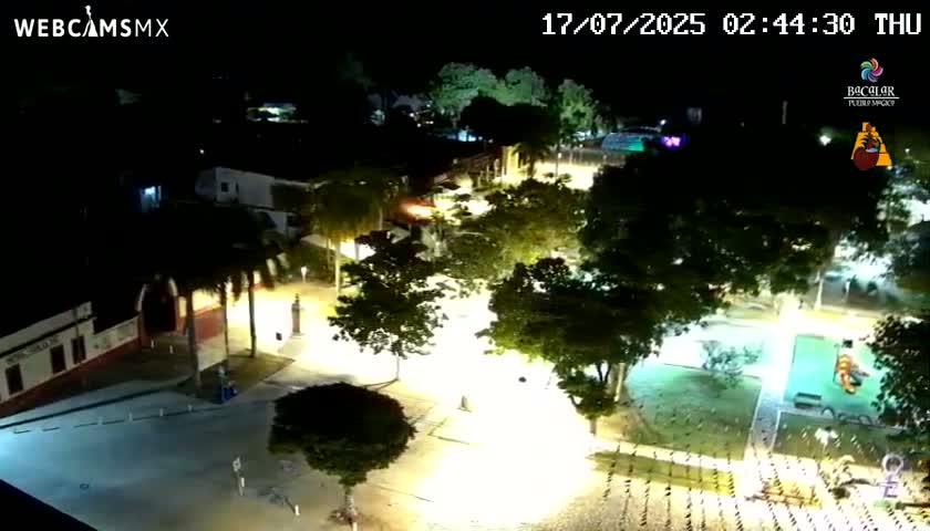A nighttime aerial view of a town square with buildings, trees, and illuminated streets.