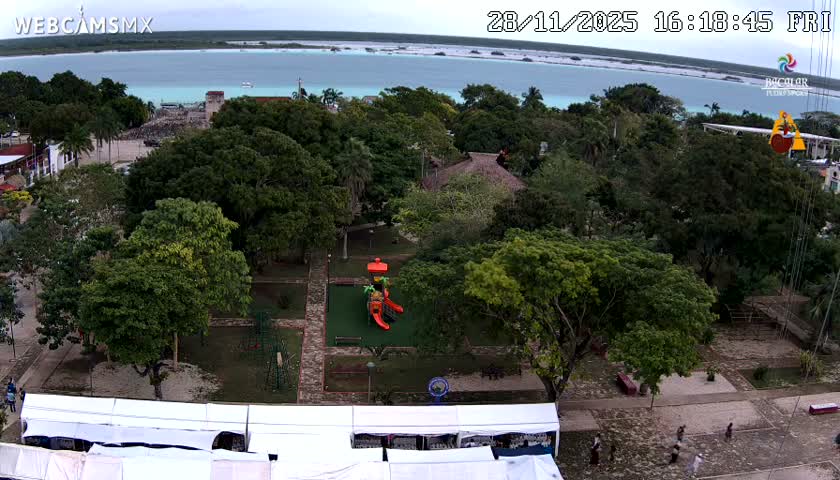 An overhead view reveals a vibrant park with lush green trees, a colorful playground, and white tents in the foreground, leading towards buildings beside a light blue bay or lagoon under a light, overcast sky.