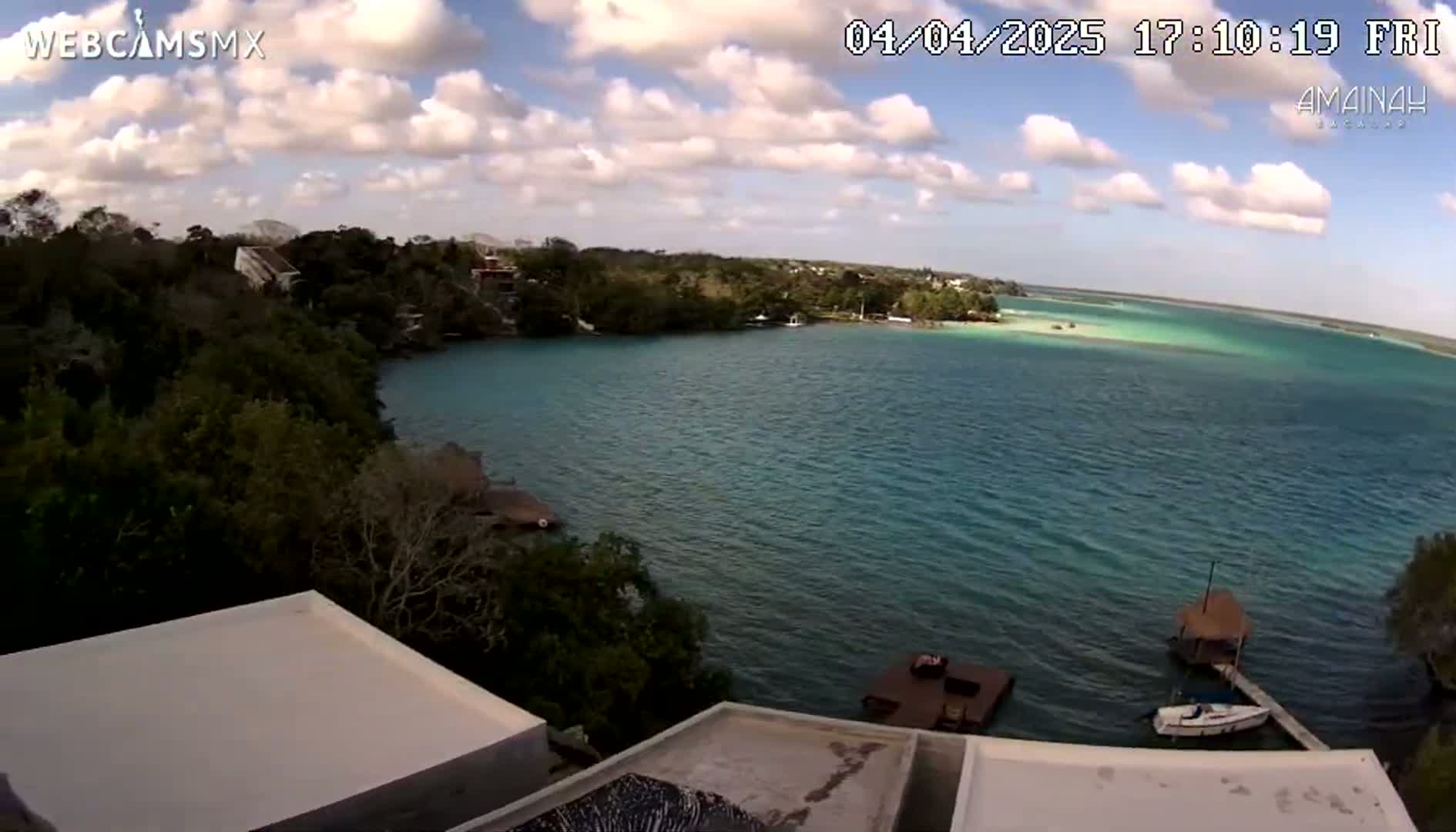 A partly cloudy day overlooking a calm, turquoise body of water, surrounded by lush green vegetation and featuring a small boat docked near a pier.