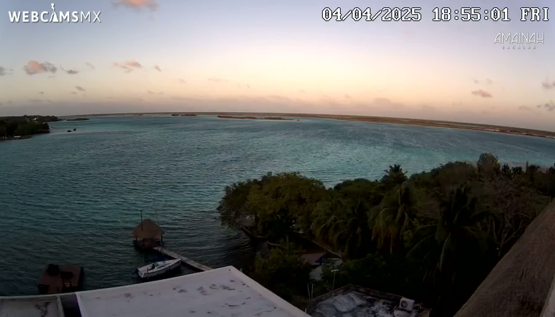 An elevated view shows a calm body of water at sunset, with a shoreline featuring lush green vegetation and a few small boats.