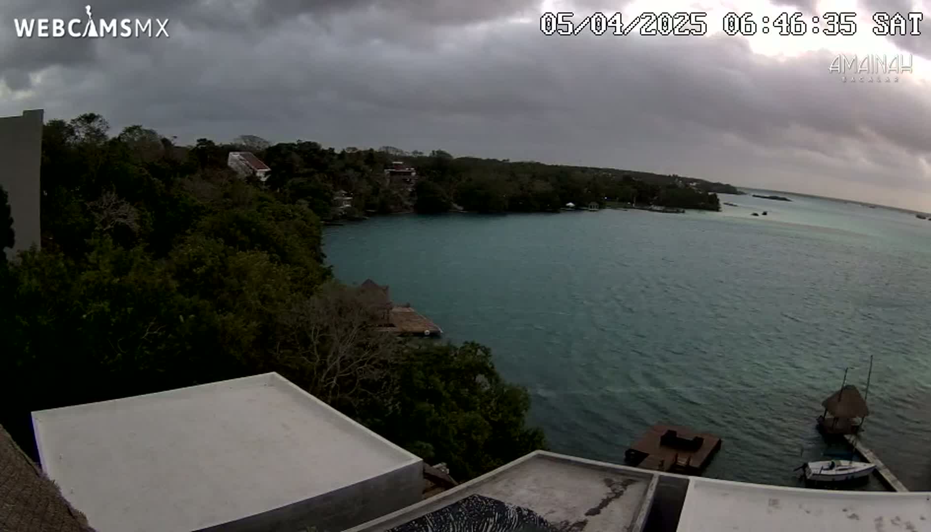 A tranquil body of water is seen under an overcast sky, viewed from a high vantage point overlooking buildings with flat roofs and lush green foliage.