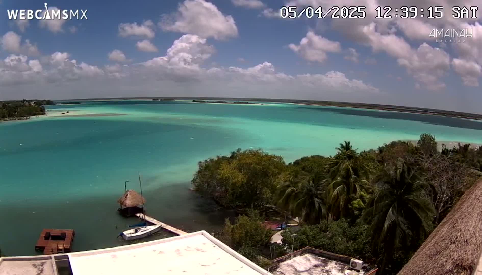 A partly cloudy sky overlooks a tranquil turquoise lagoon, bordered by lush green vegetation and a few small boats near a thatched-roof structure.