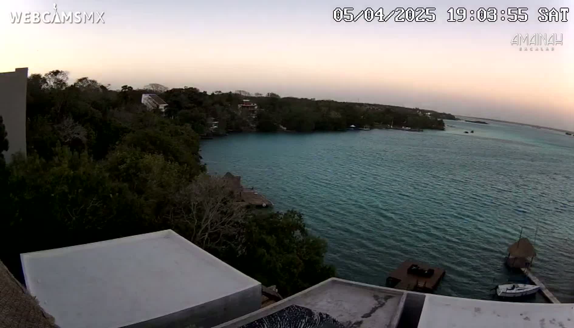 An aerial view at dusk shows a calm body of teal water surrounded by lush green vegetation, with a few buildings visible along the shoreline and a few boats moored near the shore.