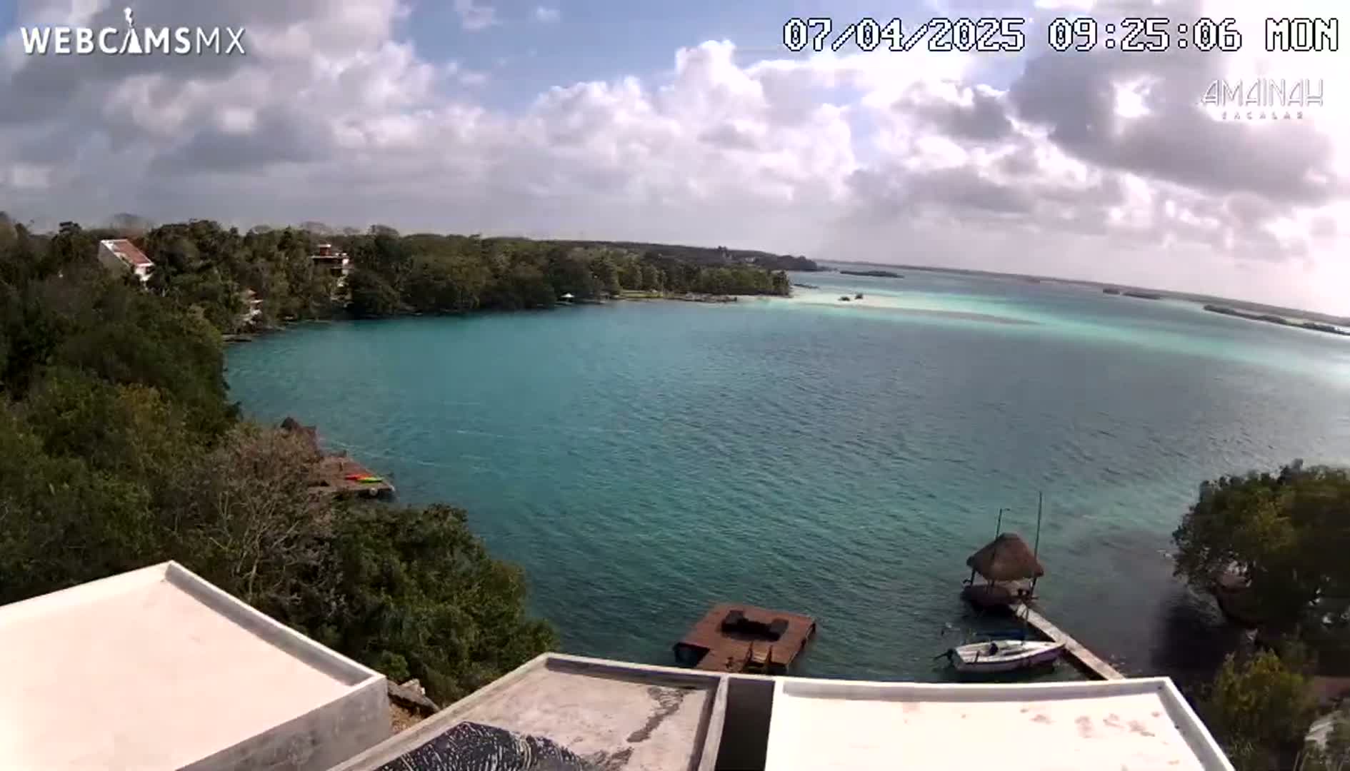 A partly cloudy day overlooking a teal-colored bay with lush green vegetation, a few boats, and a thatch-roofed structure on the shore.