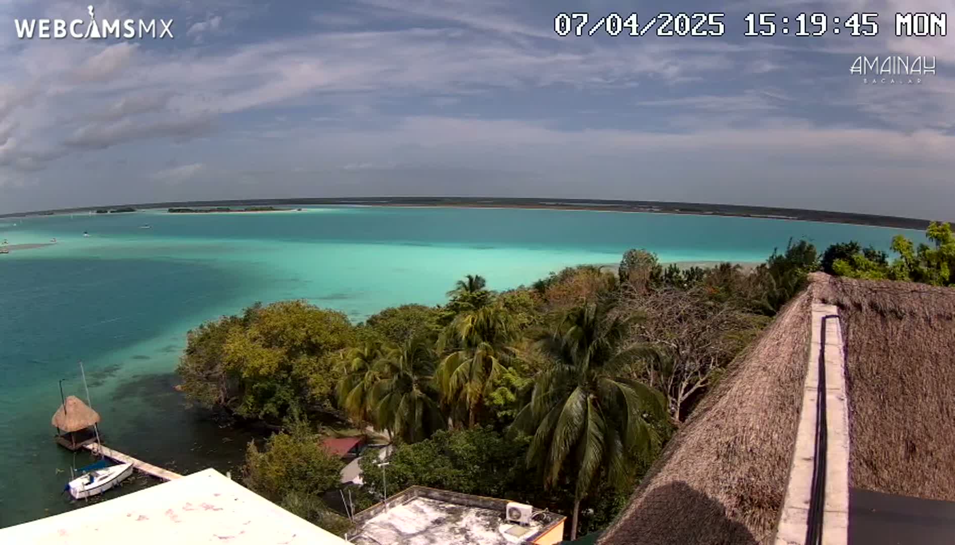 A partly cloudy day reveals a tranquil turquoise lake surrounded by lush tropical vegetation, viewed from a high vantage point near a thatched roof.