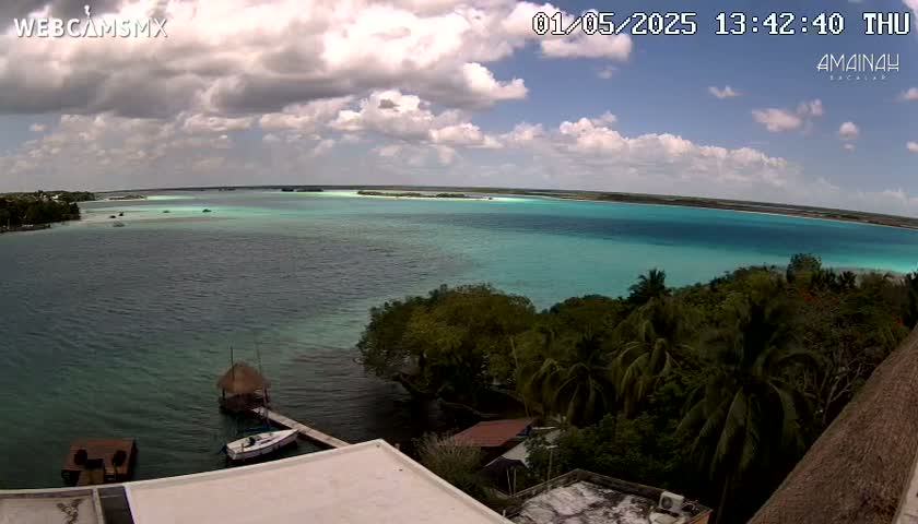 An elevated view shows a partly cloudy sky over a calm, turquoise body of water, surrounded by lush green vegetation and a few small boats near a shoreline.