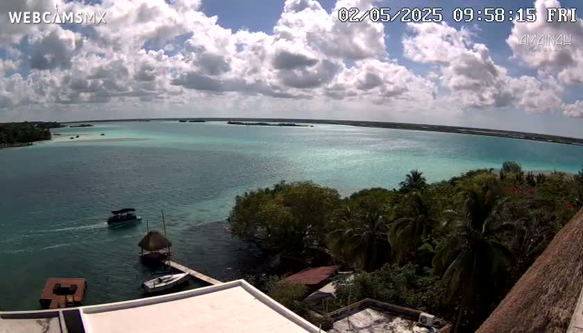 A partly cloudy day shows a tranquil turquoise bay with boats and tropical vegetation, viewed from an elevated position.