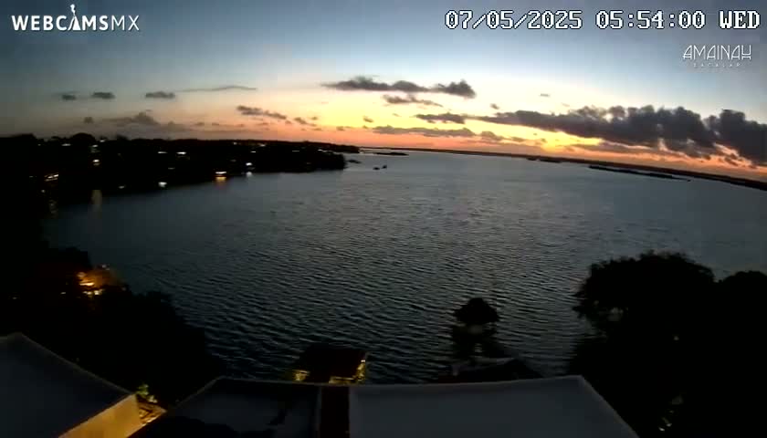 A sunset over a calm body of water, with a shoreline of dark trees and buildings visible in the distance.