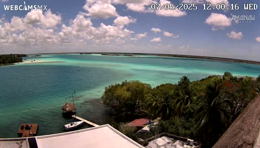A partly cloudy day reveals a tranquil turquoise lagoon, partially surrounded by lush green vegetation and a few small boats near a dock.