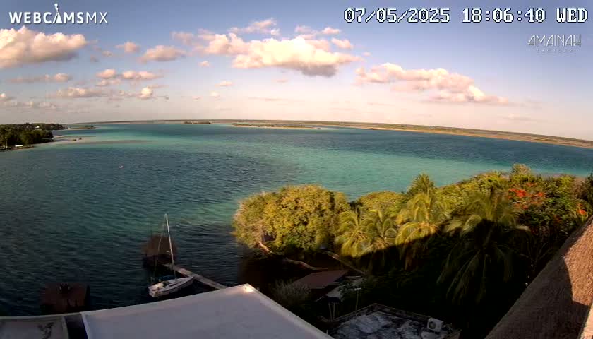 A partly cloudy afternoon view overlooking a calm, turquoise body of water, with lush green vegetation and a sailboat visible in the foreground.