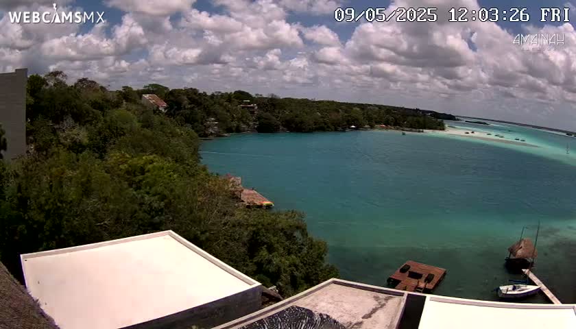 A partly cloudy day overlooks a calm, teal-colored bay surrounded by lush green vegetation and a few buildings with flat roofs.