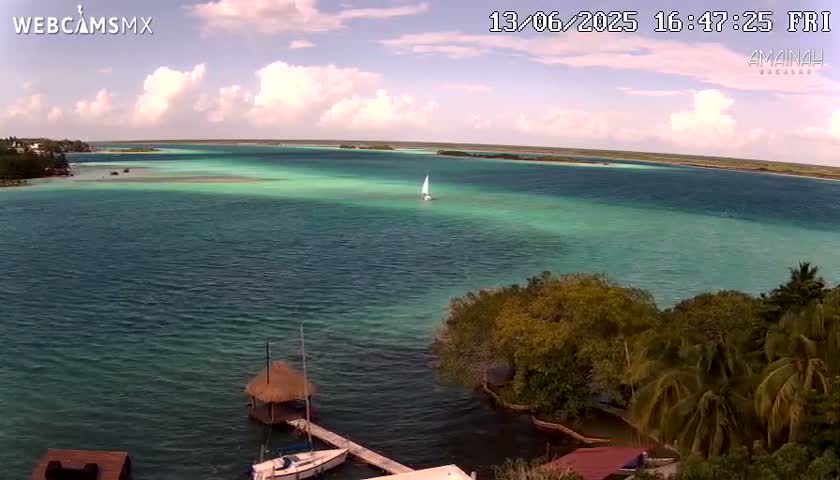 A partly cloudy sky overlooks a calm, turquoise body of water with a sailboat in the distance and a thatched-roof hut near a dock on the shore.